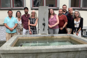 Neun Personen, davon fünf Männer und vier Frauen, posieren für ein Foto bei einem Brunnen.